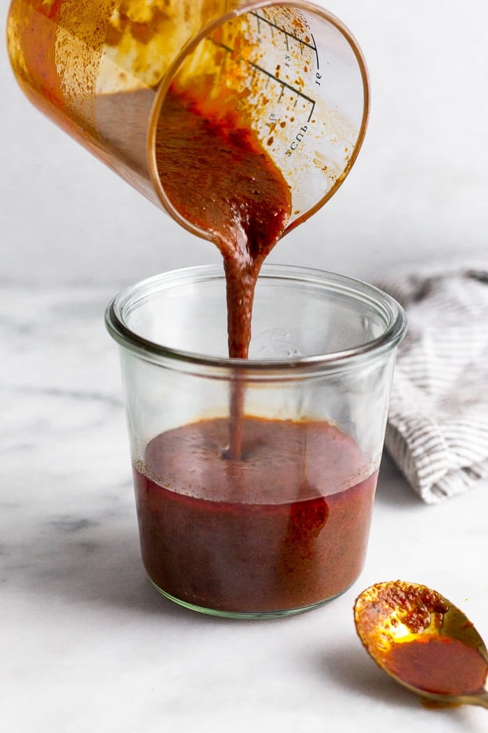 Homemade enchilada sauce being poured into a jar from a measuring cup. In front of the jar is a spoon with enchilada sauce on it.