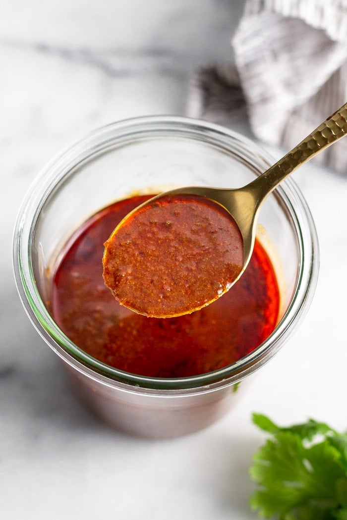 Jar of enchilada sauce with a spoonful hovering off the top. Next to the jar is cilantro and a stripped towel.