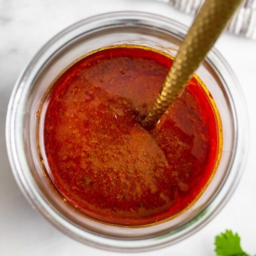 A jar of homemade red enchilada sauce with a spoon in it. Next to the jar is some cilantro and a kitchen towel.