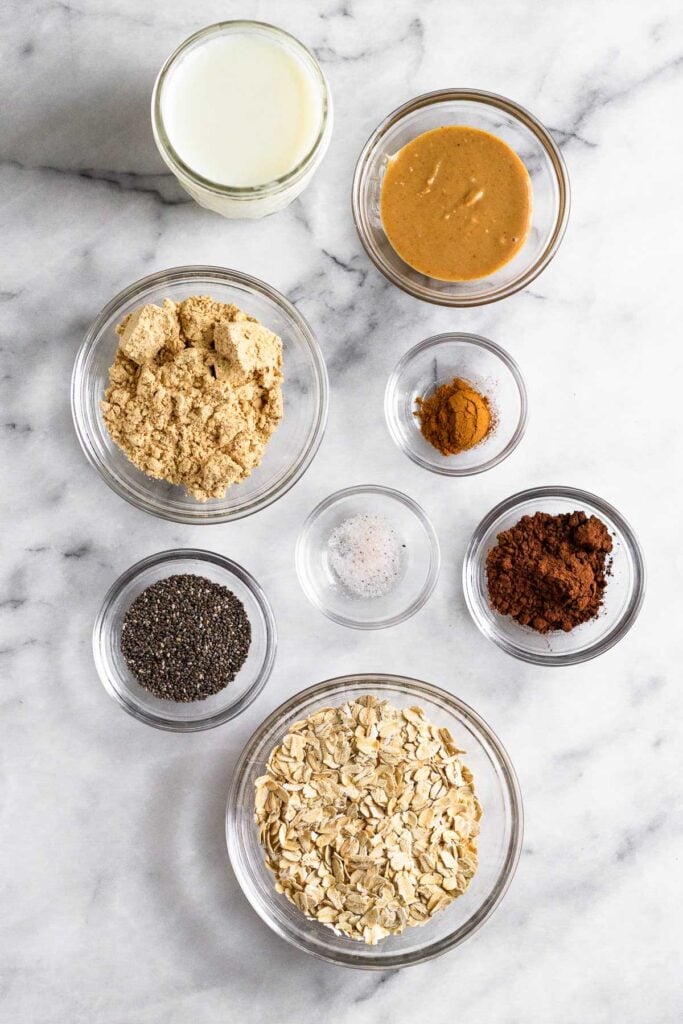 Marble counter with a jar of milk, a bowl of peanut butter, a bowl of cinnamon, a bowl of cocoa powder, a bowl of rolled oats, a bowl of chia seeds, a bowl of salt, and a bowl of protein powder.