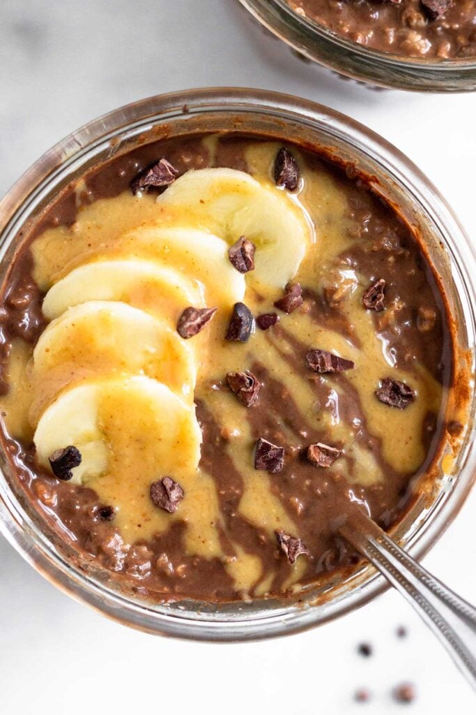 Overhead shot of a jar of chocolate overnight oats topped with sliced banana, peanut butter, and cacao nibs with a spoon in the jar.