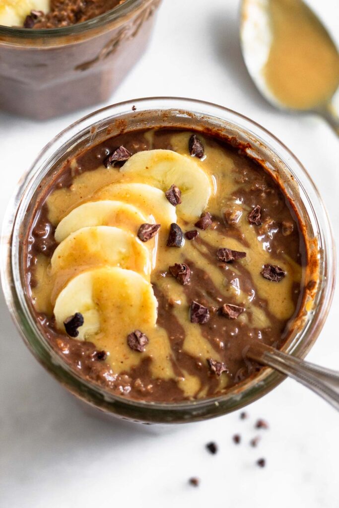Chocolate peanut butter overnight oats in a jar topped with sliced banana, peanut butter, and cacao nibs. A spoon is coming out of the jar. Behind the jar is another jar of overnight oats and a spoon with peanut butter on it.