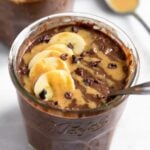 Chocolate peanut butter overnight oats in a jar topped with sliced banana, peanut butter, and cacao nibs with a spoon in the jar. Behind it is a another jar with overnight oats and a spoon with peanut butter on it.