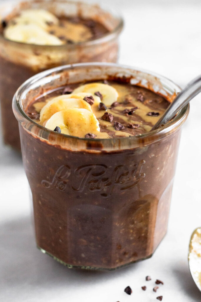 Two jars of peanut butter chocolate overnight oats, with one being behind the other. They are topped with banana, peanut butter, and cacao nibs. A spoon is in the jar in the front.
