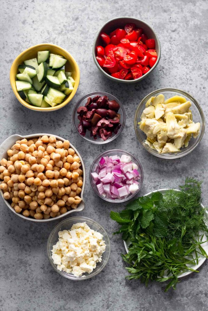 Gray counter with a bowl of diced tomatoes, a bowl of artichoke hearts, a plate of fresh herbs, a bowl of crumbled feta, a bowl of diced red onion, a bowl of chickpeas, a bowl of sliced Kalamata olives, and a bowl of diced cucumber.
