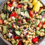 Large bowl filled with greek chickpea salad garnished with fresh herbs and two lemon wedges. Next to it is an empty jar and a bowl full of herbs.