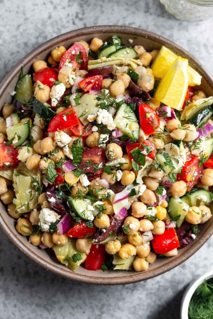 Large bowl filled with greek chickpea salad garnished with fresh herbs and two lemon wedges. Next to it is an empty jar and a bowl full of herbs.
