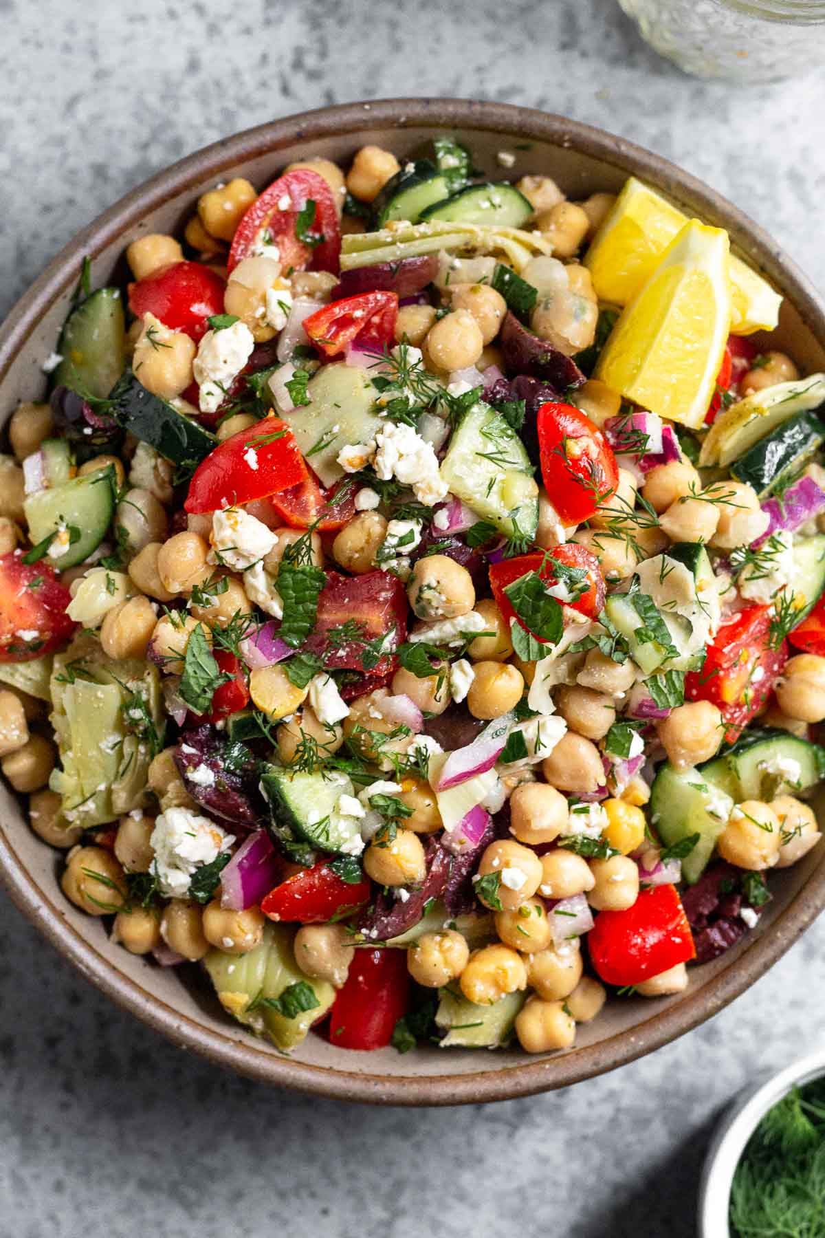 Large bowl filled with greek chickpea salad garnished with fresh herbs and two lemon wedges. Next to it is an empty jar and a bowl full of herbs.
