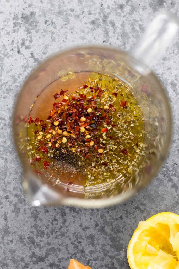 Glass measuring cup filled with red wine vinegar, olive oil, lemon, and spices. Next to it is half a juiced lemon.