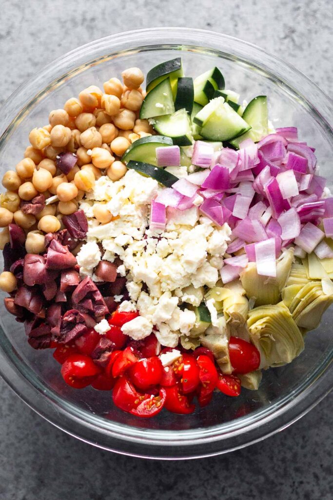 Large glass bowl filled with chickpeas, chopped cucumbers, diced red onion, artichoke hearts, diced tomatoes, Kalamata olives, and crumbled feta.