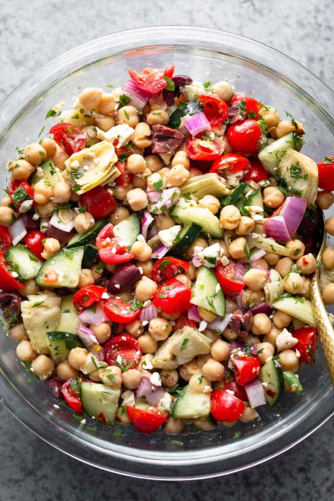 Mediterranean chicken salad with tomatoes, cucumbers, olives, artichoke hearts, feta, and herbs tossed with dressing in a glass large bowl.