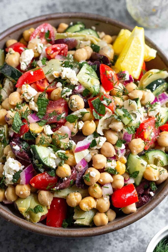 Large bowl filled with Mediterranean chickpea salad garnished with fresh herbs and two lemon wedges. Next to it is an empty jar and a bowl full of herbs.