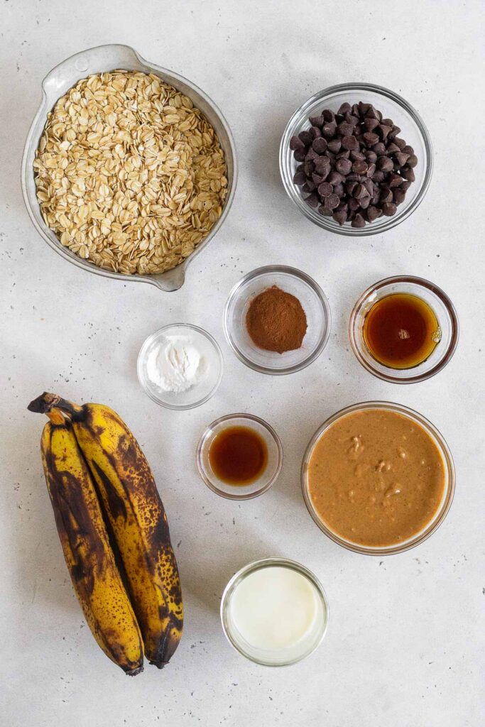 White counter with a bowl of rolled oats, a bowl of chocolate chips, a bowl of cinnamon, a bowl of maple syrup, a bowl of peanut butter, a jar of milk, a bowl of vanilla extract, two brown bananas, and a bowl of baking powder.