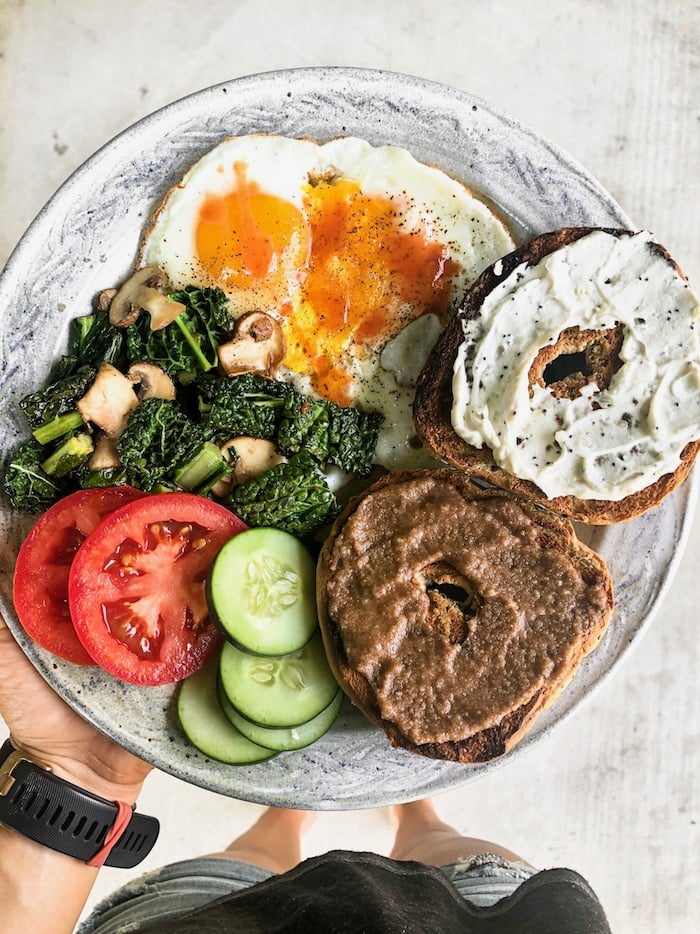 Plate of a 2 fried eggs, veggies, and a bagel with half with cream cheese and half with nut butter.