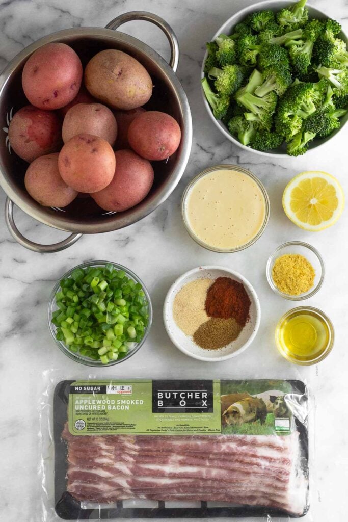 White marble counter with a bowl of broccoli florets, a bowl of mayo, half a lemon, a bowl of nutritional yeast, a bowl of olive oil, a pack of bacon, a bowl of spices, a bowl of sliced green onions, and a colander of red potatoes.