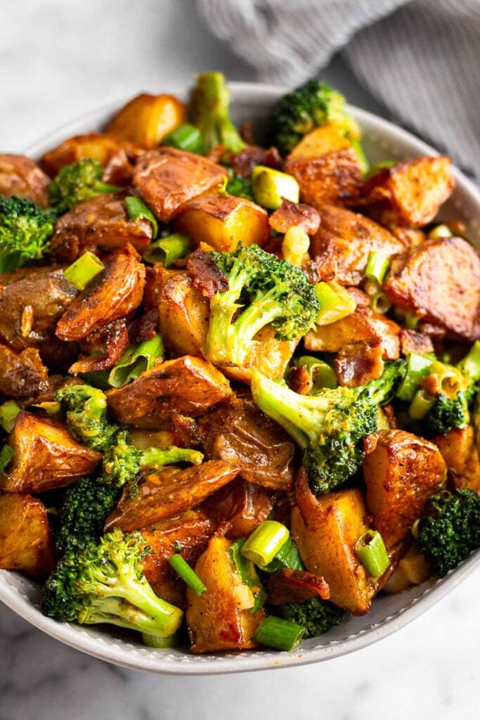 Close up of a bowl with potato salad with bacon and broccoli in it. Behind the bowl off in the distance is a striped kitchen towel.