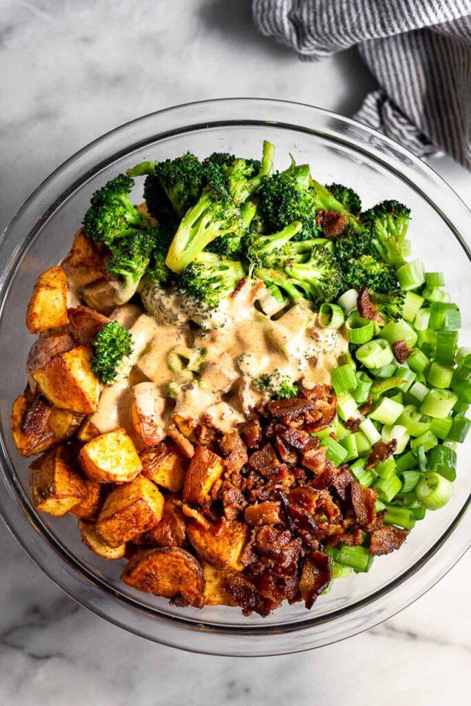 Large glass bowl filled with roasted potatoes, sautéed broccoli, crumbled bacon, sliced green onions, and a spicy mayo poured on top.