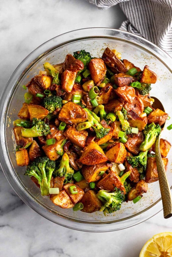 Large glass bowl filled with roasted potato salad with broccoli and bacon with a spoon in it. Next to the bowl is half a lemon and a striped towel.