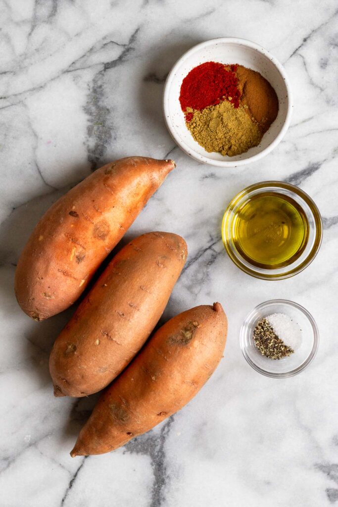 White marble counter top with a bowl of spices, a bowl of oil, a bowl of salt and pepper, and 3 sweet potatoes.