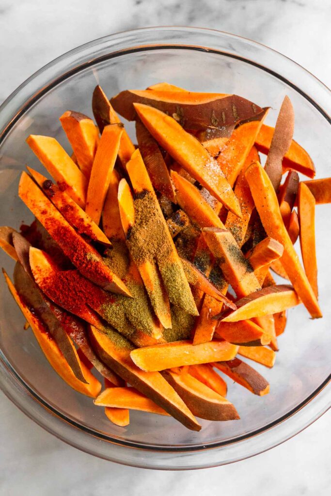 Overhead shot of raw sweet potatoes cut into french fries sprinkled with spices in a large bowl.