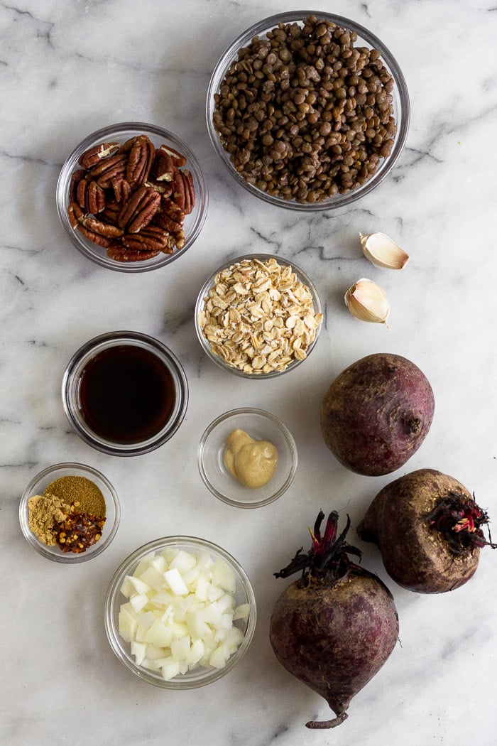 white counter top with a bowl of cooked lentils, whole beets, diced onion, oats, spices, coconut animos, mustard, garlic, and pecans.