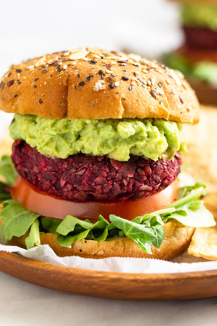 Close up of a beet burger with lettuce, tomato, and avocado. It is on a wooden plate with another burger in the distance behind it.