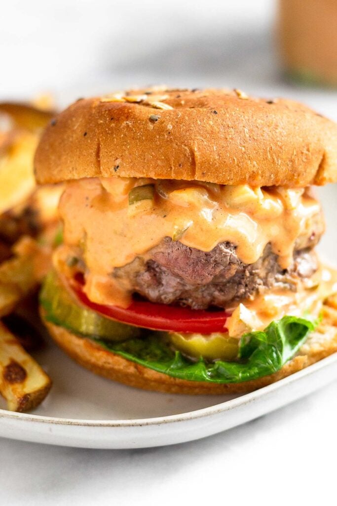 Close up of a hamburger on a bun with lettuce, tomato, and pickles topped with special sauce that is dripping down the burger.