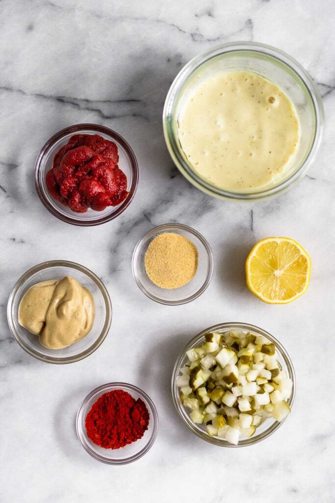 White marble counter with a jar of mayo, half a lemon, a bowl of diced pickles, a bowl of paprika, a bowl of mustard, a bowl of garlic powder, and a bowl of tomato paste.