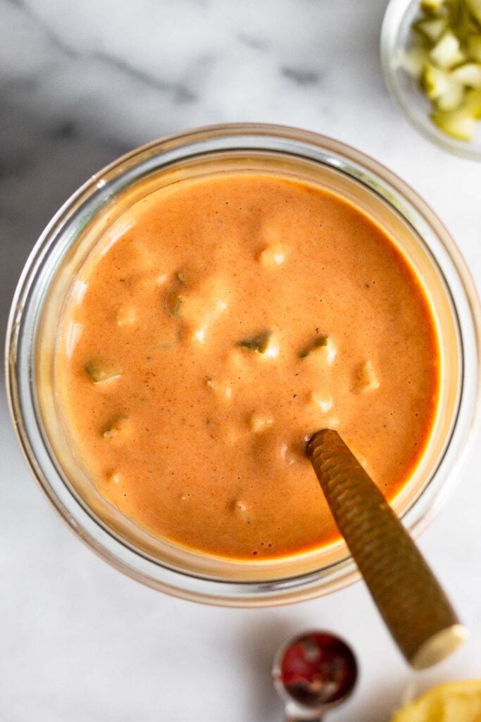 Overhead shot of a large jar with burger sauce and a spoon in it. Around it is a bowl of diced pickles, half a lemon, and a teaspoon of tomato paste.