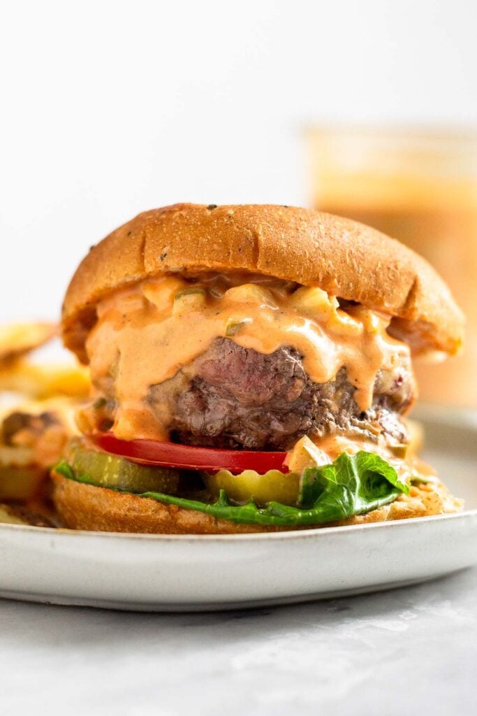 A burger on a plate with lettuce, tomato, and pickles, and topped with burger sauce that is dripping down the burger. Behind it is a jar of more burger sauce.