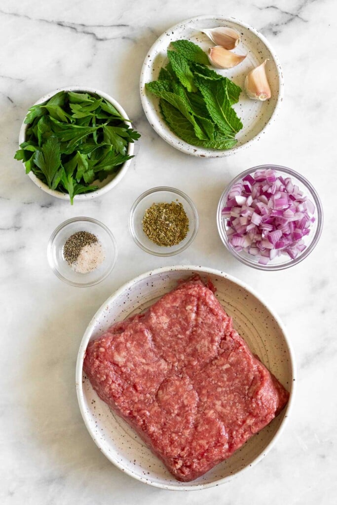 White marble counter with a small plate with fresh mint and 3 garlic cloves on it, a bowl of diced red onion, a plate of raw ground lamb, a bowl of spices, a bowl of salt and pepper, and a bowl of fresh parsley.