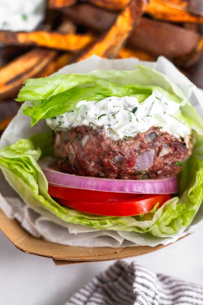 Close up of a greek burger wrapped in lettuce with sliced tomato, red onion, and tzatziki sauce. Behind the burger are sweet potato fries and more tzatziki sauce.