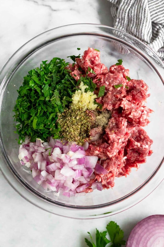 Large glass bowl of ground lamb, diced red onion, chopped fresh herbs, minced garlic, spices, and salt and pepper. Next to the bowl is a striped towel, half a red onion, and some fresh parsley.