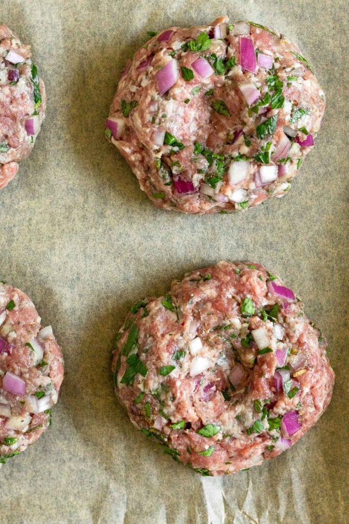 Raw burgers with onion and fresh herbs in them on a piece of parchment paper.