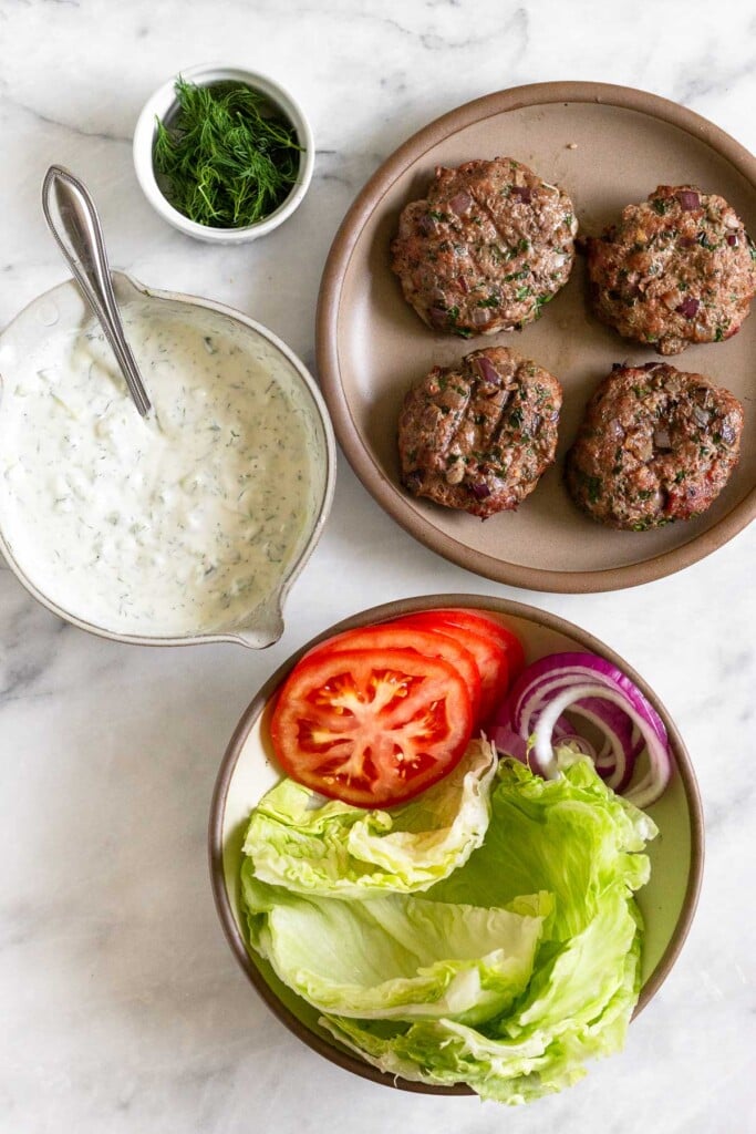 White marble counter with a plate of 4 burgers, a bowl of lettuce, sliced tomatoes, and sliced onions, a bowl of tzatziki sauce, and a small dish of fresh dill.
