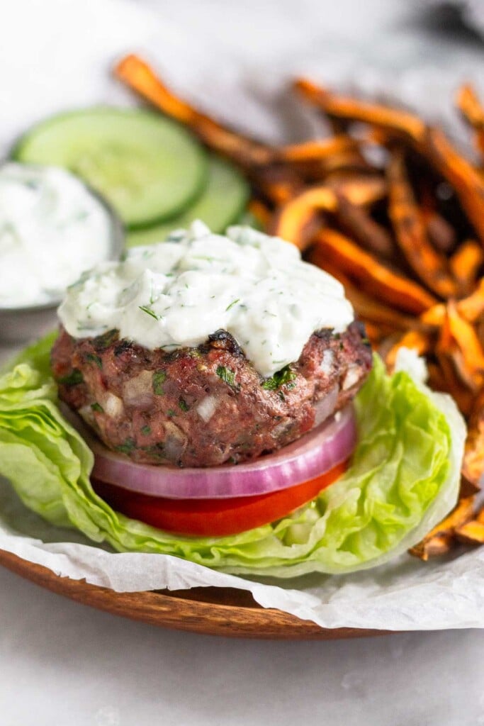 A plate with a mediterranean burger with tomato, red onion, and tzatziki sauce sitting on a lettuce cup. Behind the burger on the plate are sweet potato fries, sliced cucumbers, and a dish of tzatziki sauce.
