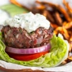 Lamb burger sitting on a lettuce cup with sliced tomato, red onion, and topped with tzatziki sauce. Behind the burger are sweet potatoes fries, sliced cucumbers, and a dish of tzatziki sauce.