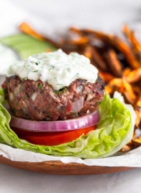 Lamb burger sitting on a lettuce cup with sliced tomato, red onion, and topped with tzatziki sauce. Behind the burger are sweet potatoes fries, sliced cucumbers, and a dish of tzatziki sauce.