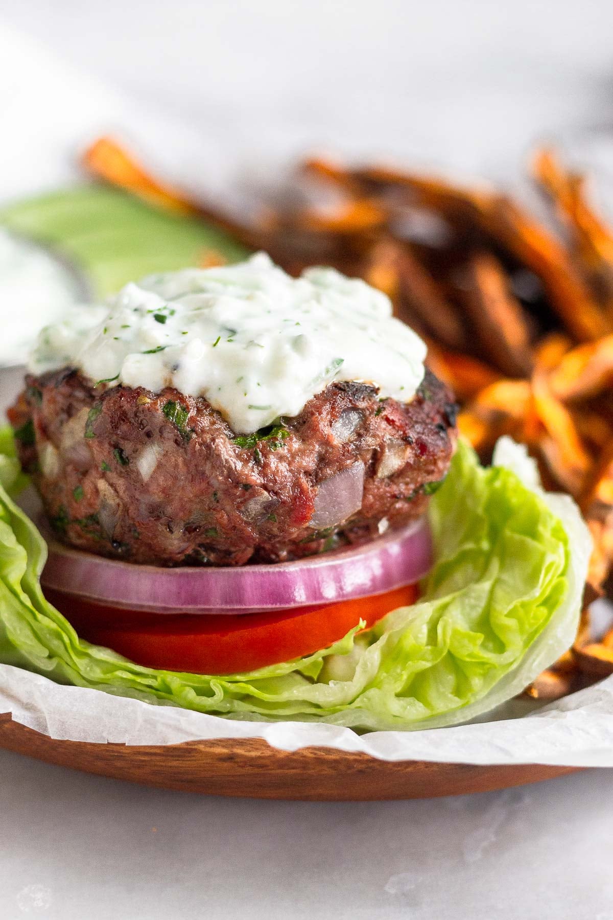Lamb burger sitting on a lettuce cup with sliced tomato, red onion, and topped with tzatziki sauce. Behind the burger are sweet potatoes fries, sliced cucumbers, and a dish of tzatziki sauce.