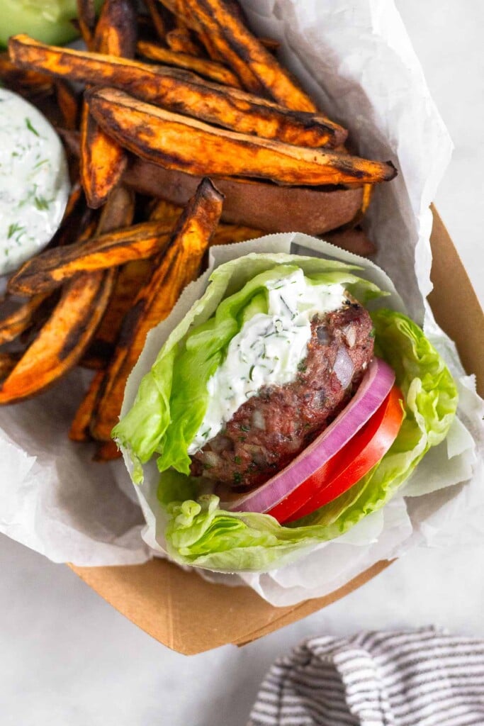 Cardboard dish filled with a lettuce wrapped lamb burger with lettuce and red onion topped with tzatziki sauce, sweet potato fires, and a dish of tzatziki sauce.
