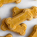 Frozen pumpkin peanut butter dog treats spread out on a white counter top with some stacked on top of each other.