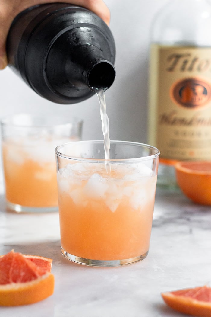Healthy cocktail recipe being poured from a shaker into a glass. Behind it is another glass and a bottle of vodka.