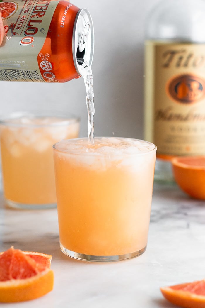 Grapefruit cocktail recipe being topped off with sparkling water. Around it is grapefruit wedges, another filled glass, half a grapefruit, and a bottle of vodka.