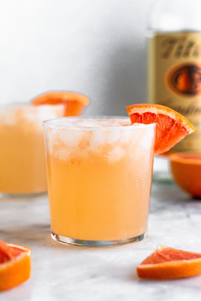 Grapefruit crush with a grapefruit wedge on the rim of the glass. Behind it is another drink, half a grapefruit, and a bottle of vodka.