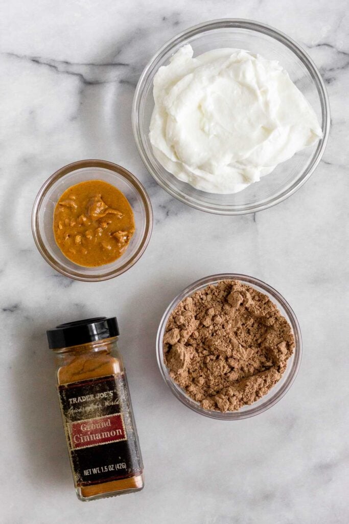Marble counter with a bowl of greek yogurt, a bowl of chocolate protein powder, a bowl of peanut butter, and a jar of cinnamon.