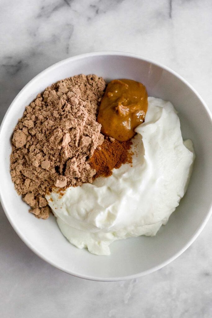 A white bowl filled with yogurt, chocolate protein powder, peanut butter, and cinnamon before it is mixed together.