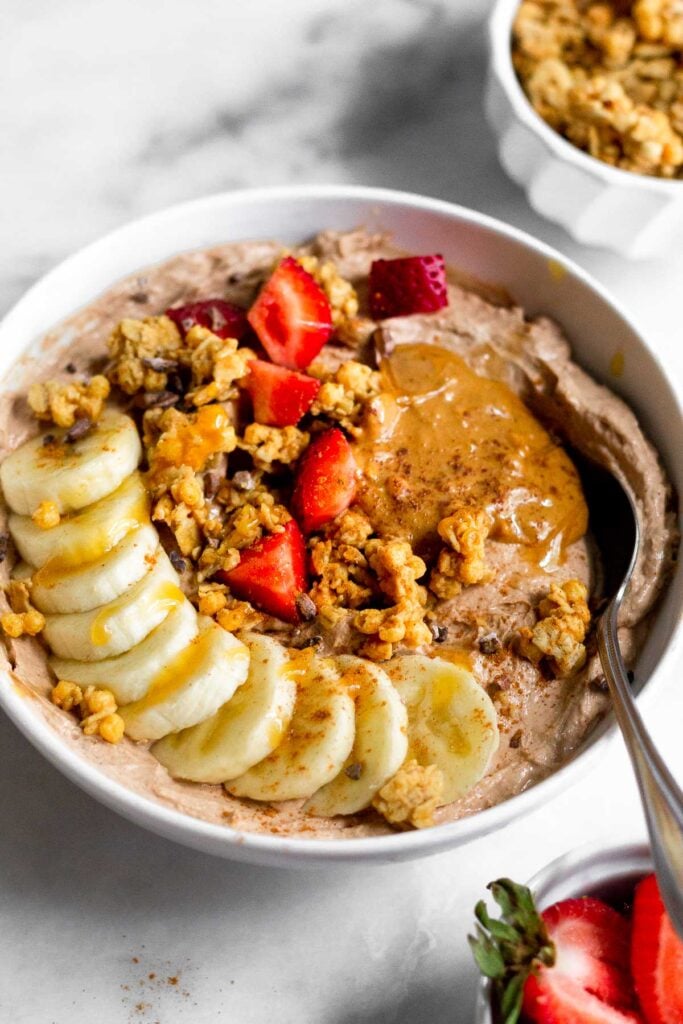 Chocolate peanut butter greek yogurt bowl topped with bananas, strawberries, granola, peanut butter, and honey. In front of it is a bowl of strawberries and behind it is a bowl of granola.