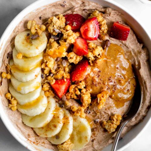 Protein yogurt bowl topped banana, granola, strawberries, peanut butter, and honey. A spoon is coming out of the bowl. Next to the bowl is a bowl of strawberries and a bowl of granola.