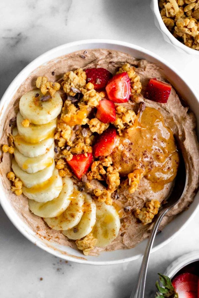 Protein yogurt bowl topped banana, granola, strawberries, peanut butter, and honey. A spoon is coming out of the bowl. Next to the bowl is a bowl of strawberries and a bowl of granola.