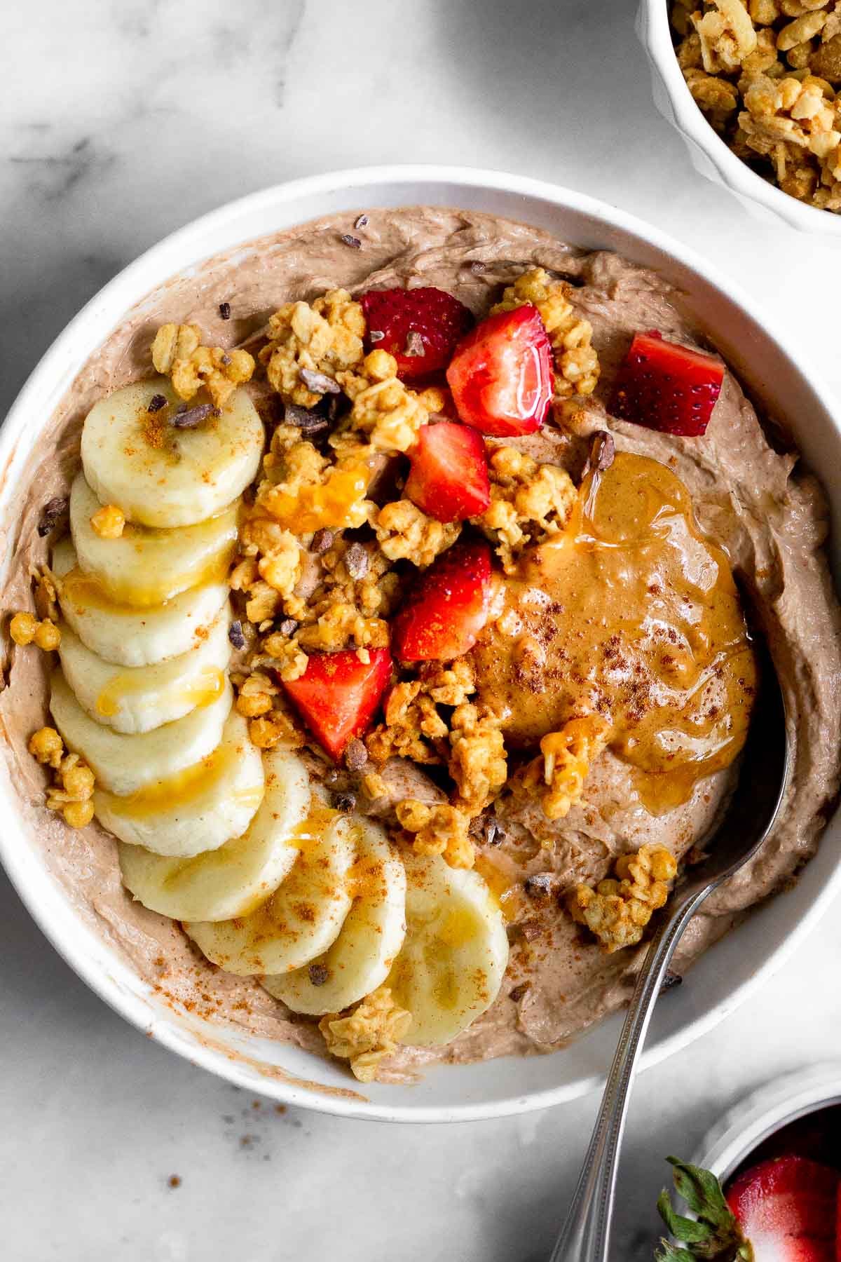 Protein yogurt bowl topped banana, granola, strawberries, peanut butter, and honey. A spoon is coming out of the bowl. Next to the bowl is a bowl of strawberries and a bowl of granola.